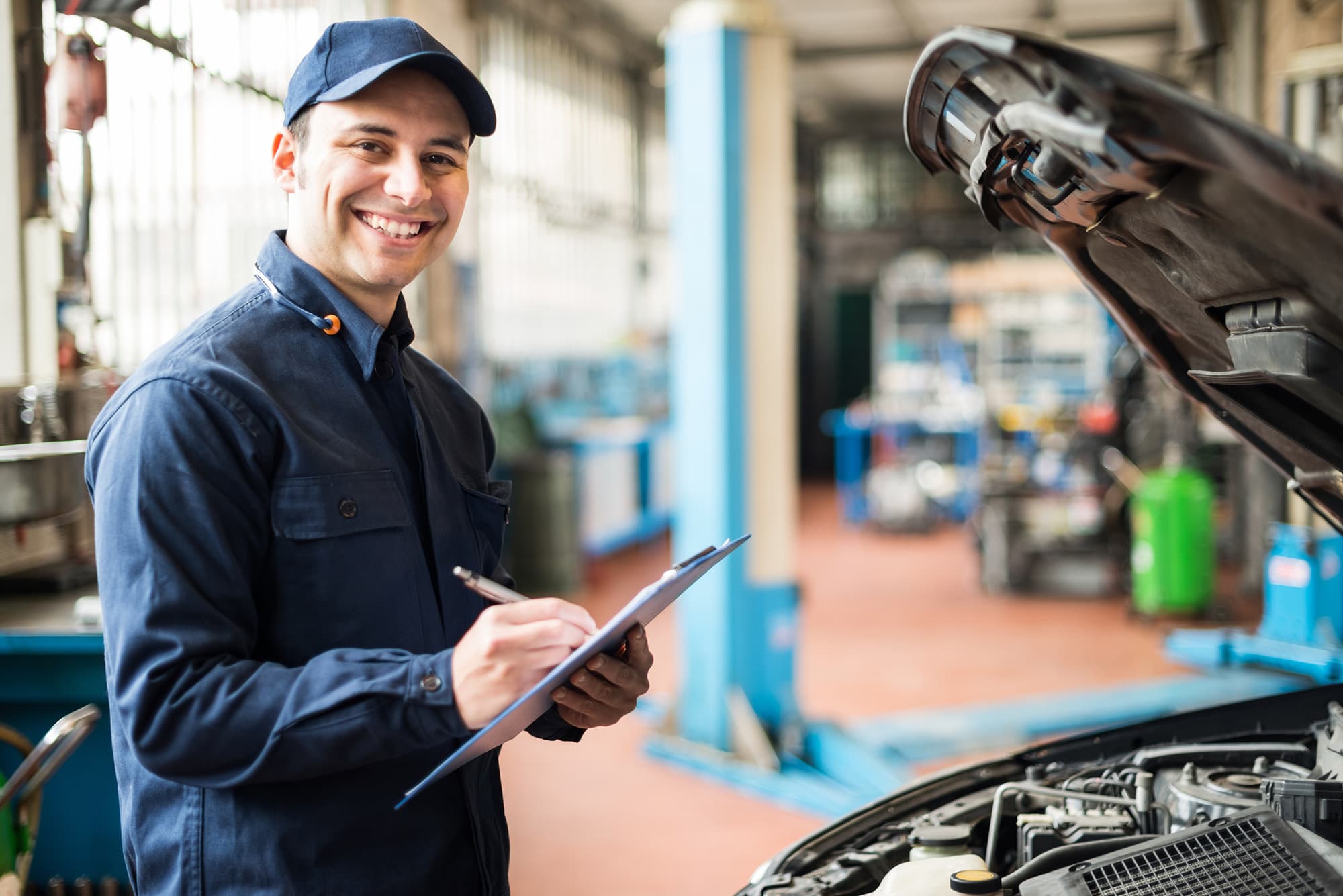 Smiling mechanic in a blue cap and coveralls writing on a clipboard beside a car with its hood raised in an auto repair shop