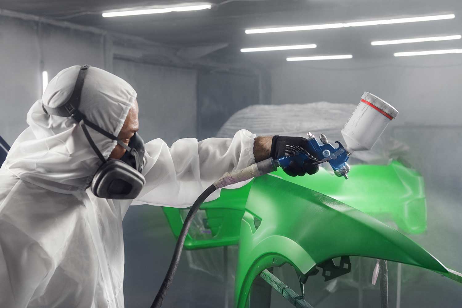 Painter in a white protective suit and respirator spraying a green car fender with a spray gun inside a misty paint booth