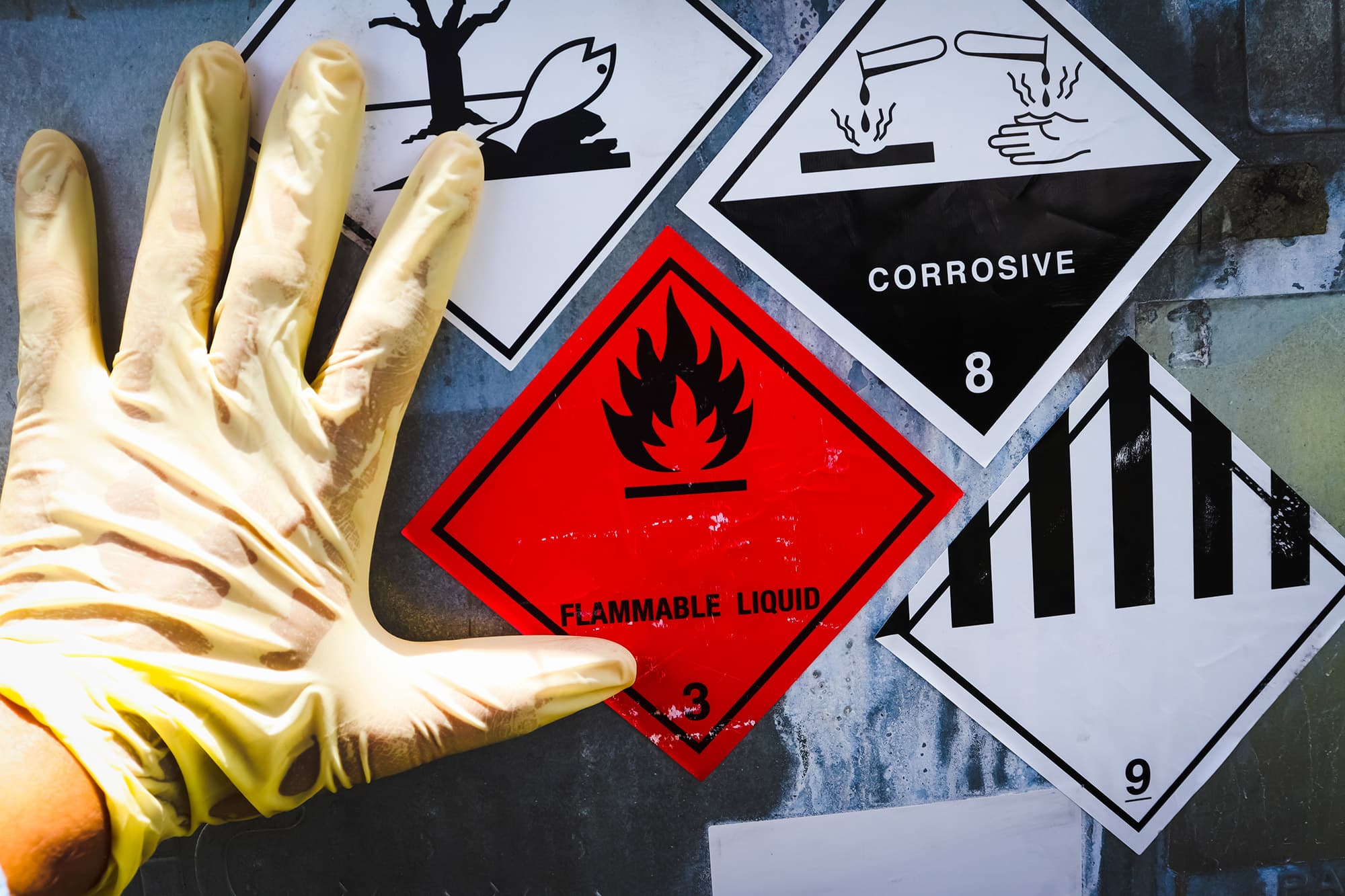 Gloved hand resting beside hazard placards, including a red Flammable Liquid diamond and a black and white Corrosive label