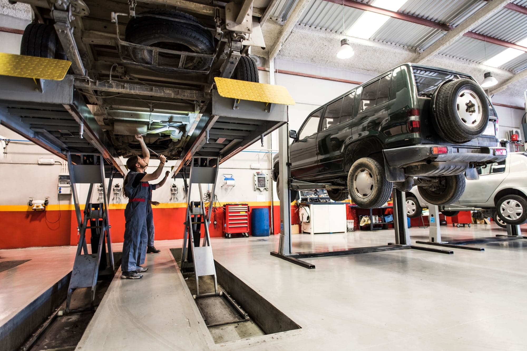 Workers beneath a vehicle raised on a service lift, with other vehicles elevated on lifts in an automotive repair shop.
