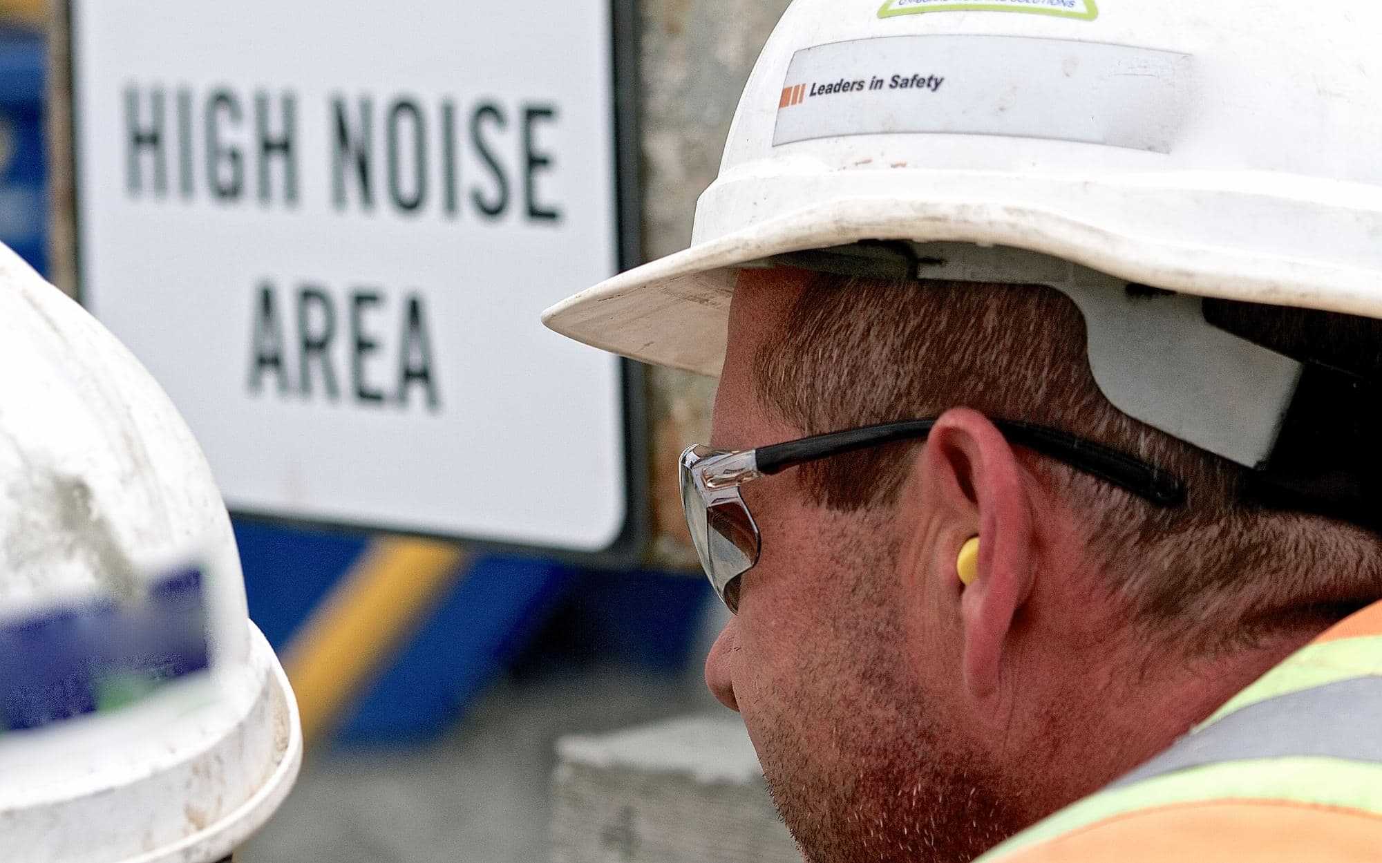 Worker wearing a hard hat, safety glasses, and an earplug near a high-noise-area sign.
