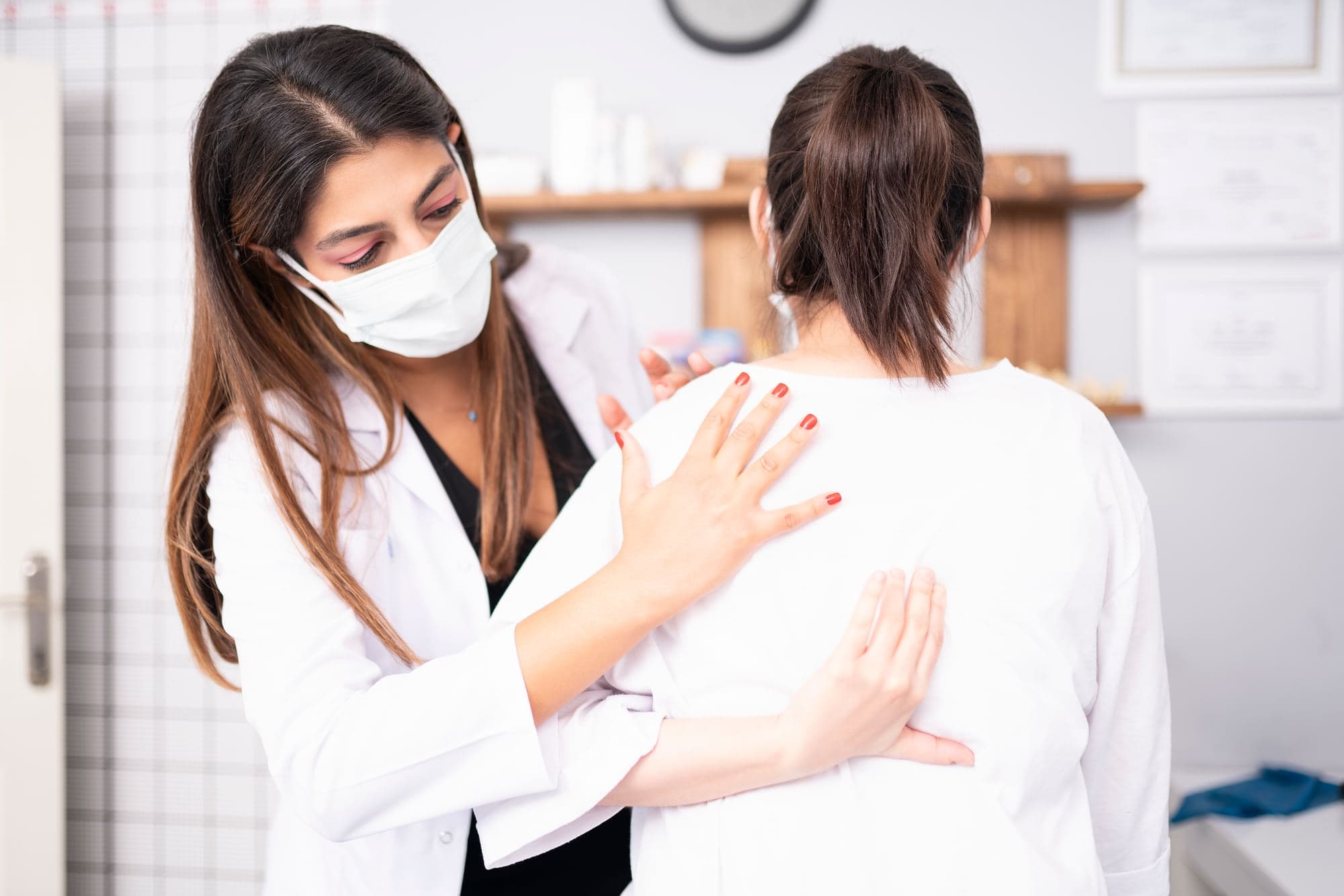 Person in a white coat and face mask placing hands on another person's shoulder and upper back.