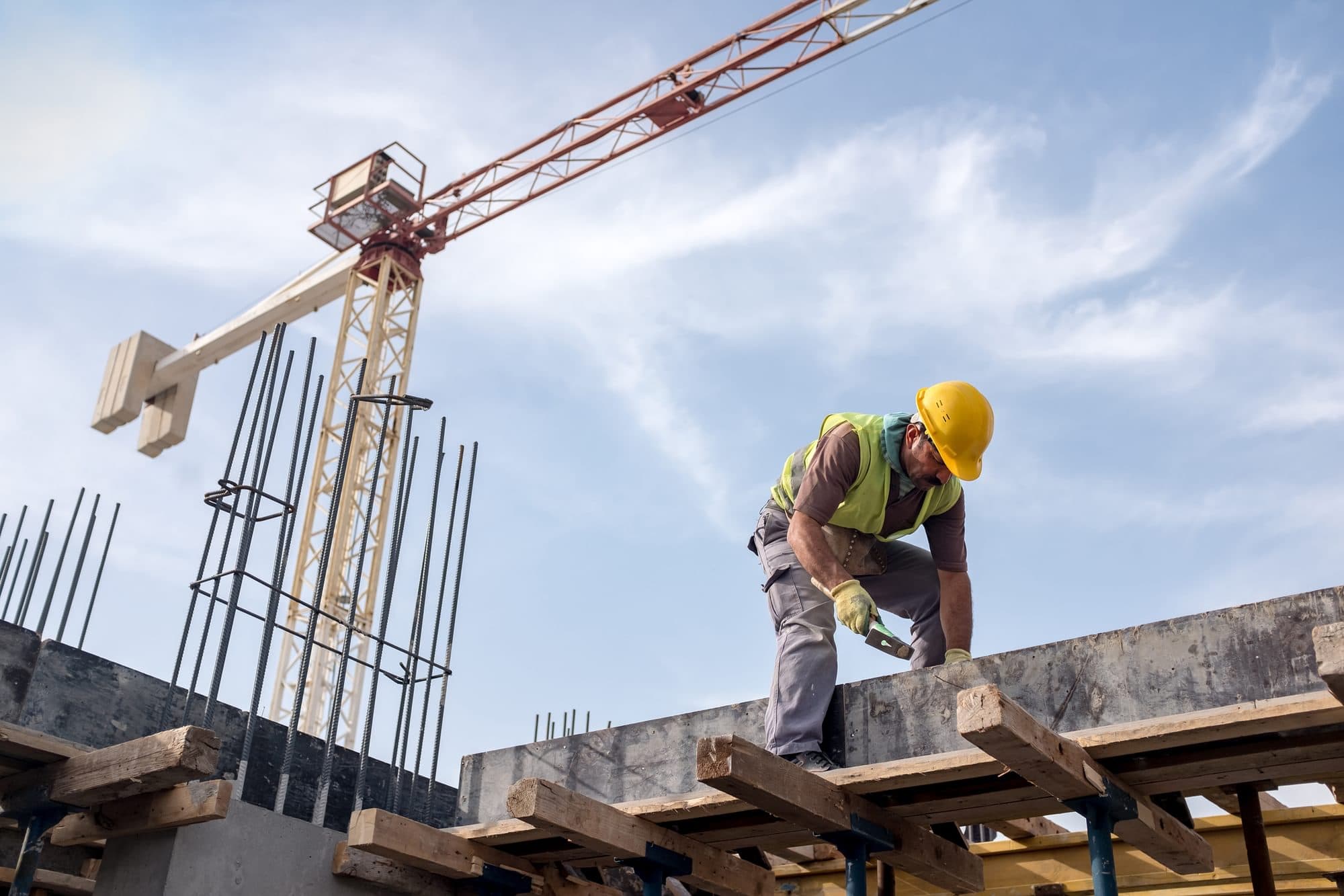 Construction worker wearing a hard hat and high-visibility vest working on concrete formwork with a tower crane in the background.