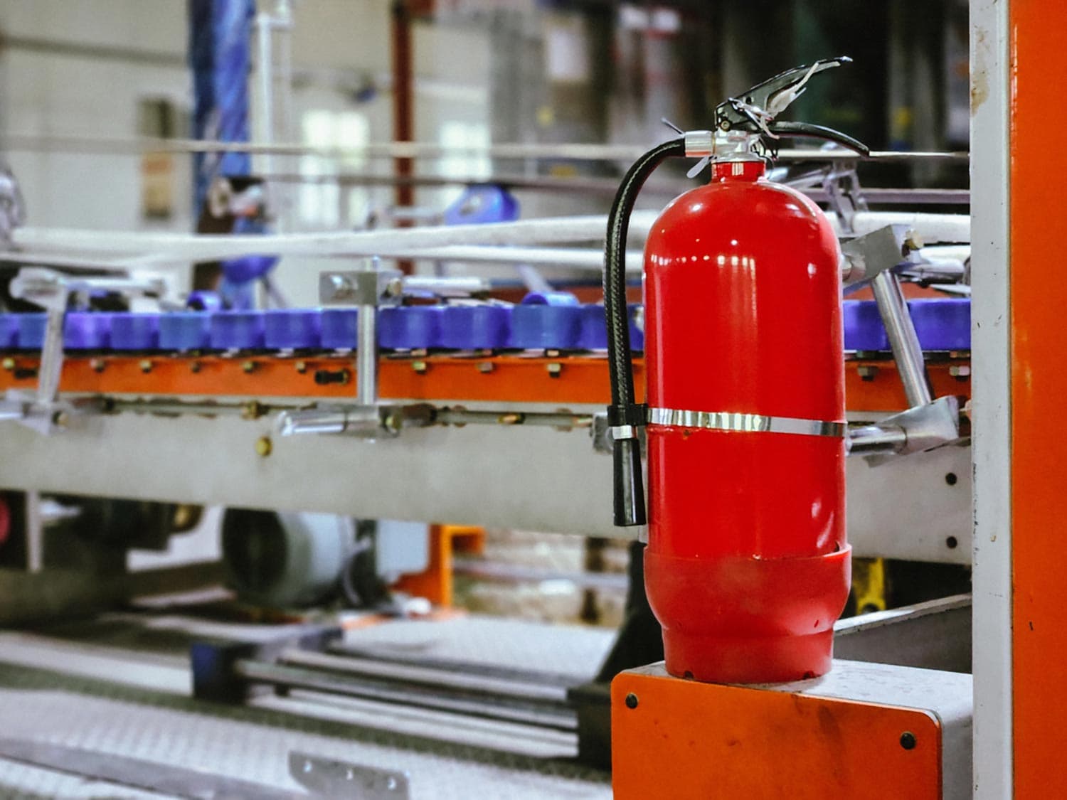 Red portable fire extinguisher mounted on a post beside a conveyor line in an industrial plant