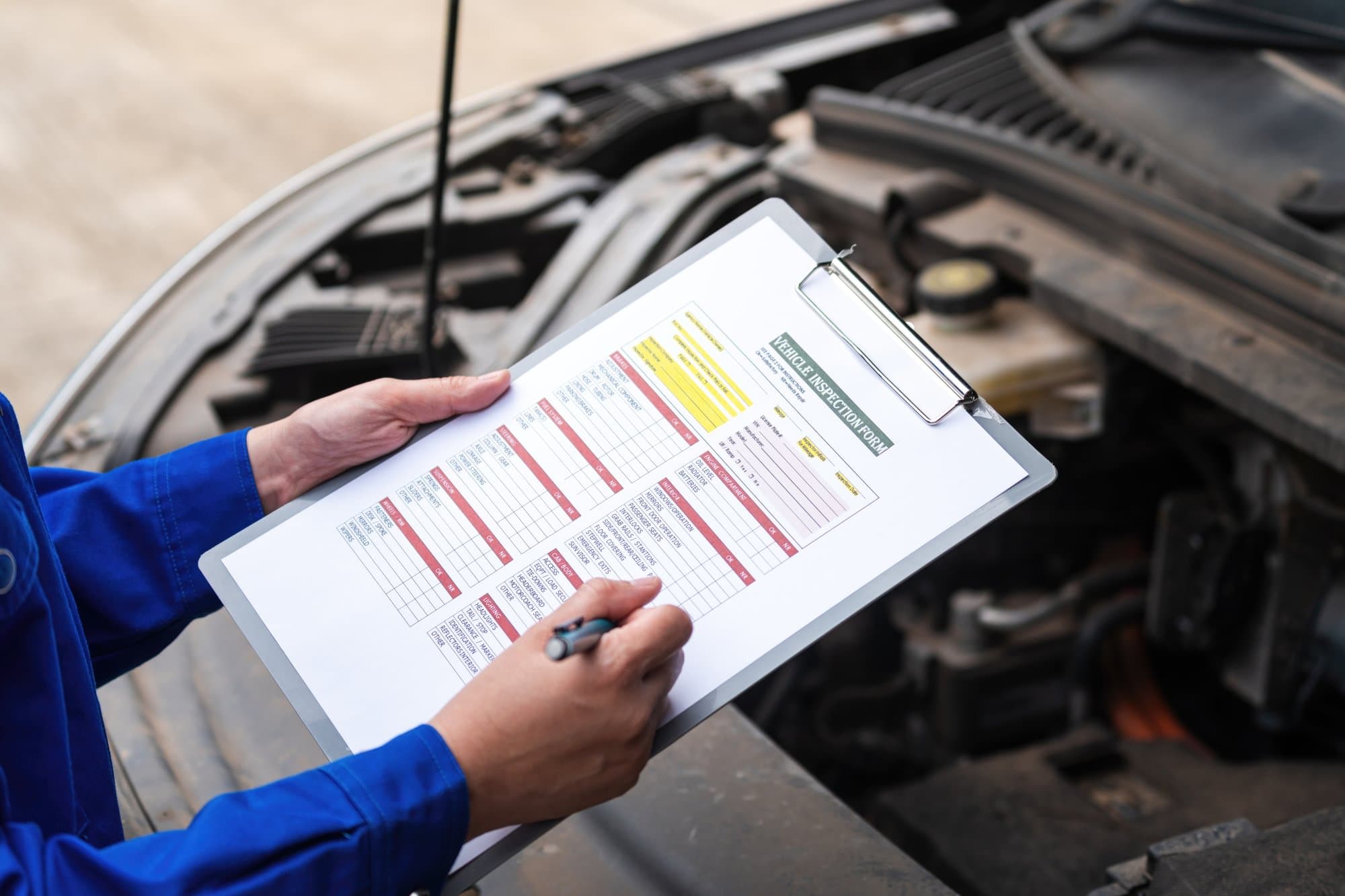 Person completing a vehicle inspection form beside an open vehicle engine compartment.
