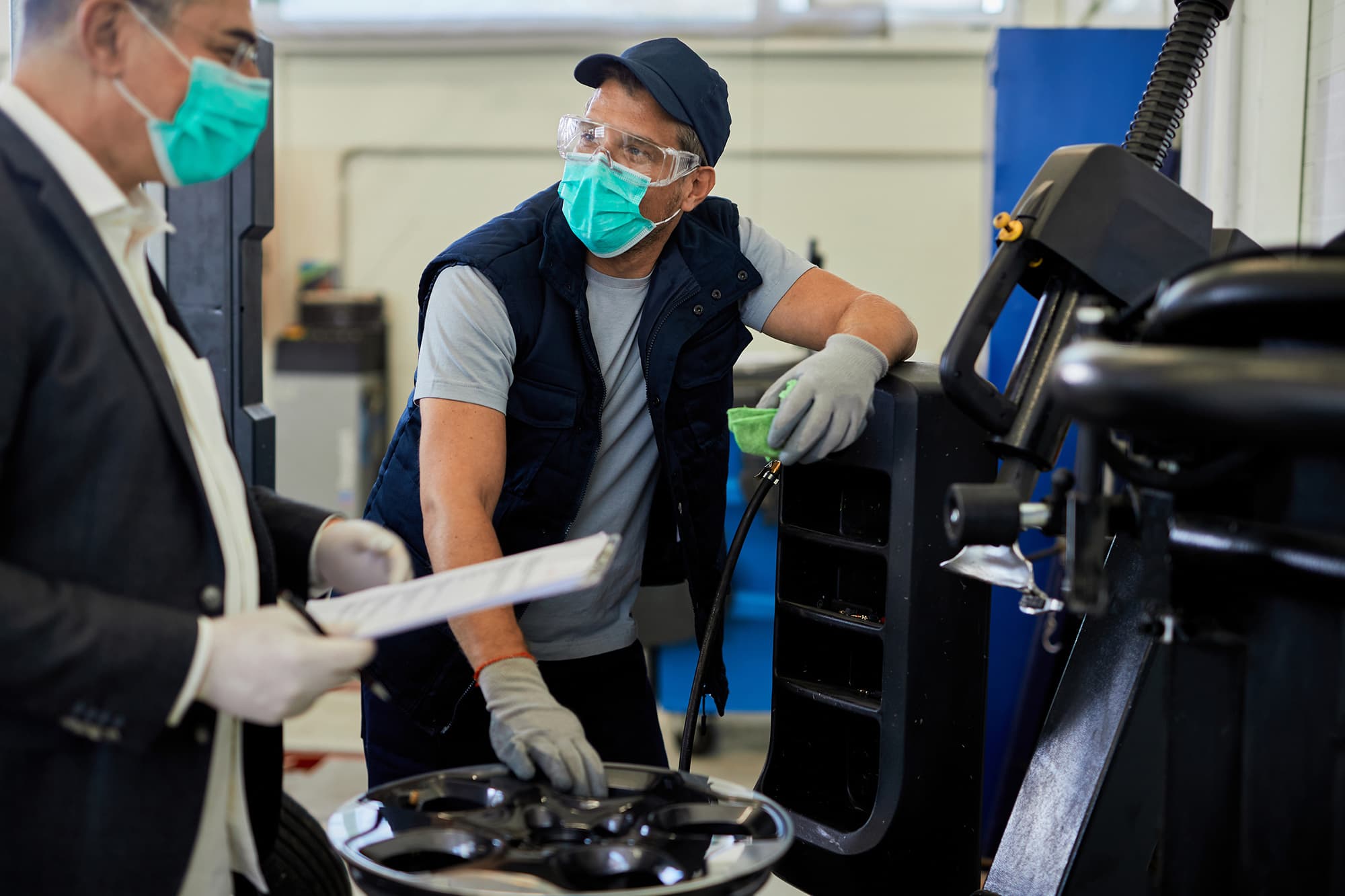 Auto mechanic in a surgical mask, safety glasses and gloves wiping down tire-service equipment while a masked manager holds a clipboard