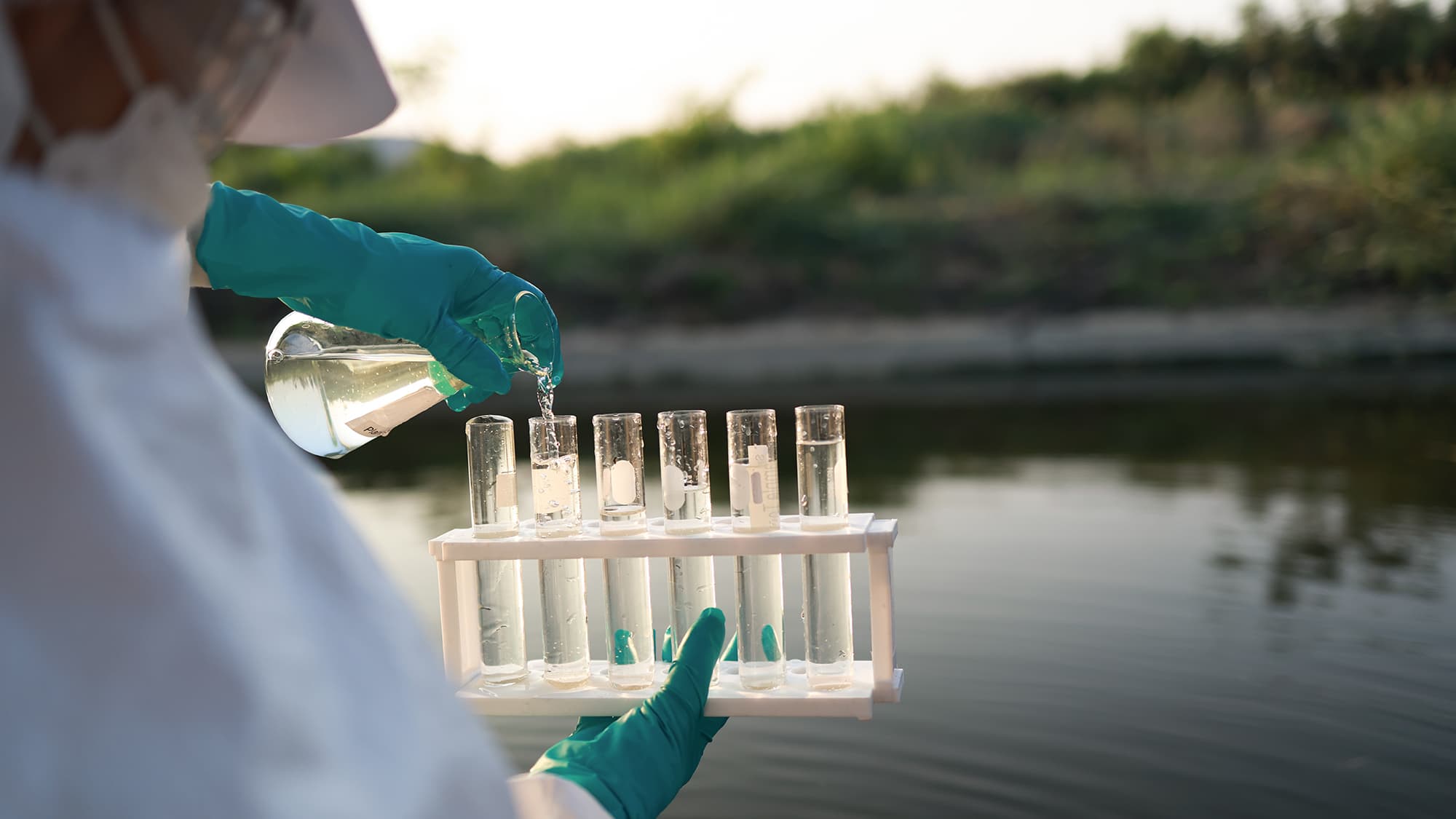 Gloved technician in protective clothing pouring a water sample from a flask into labeled test tubes beside a pond
