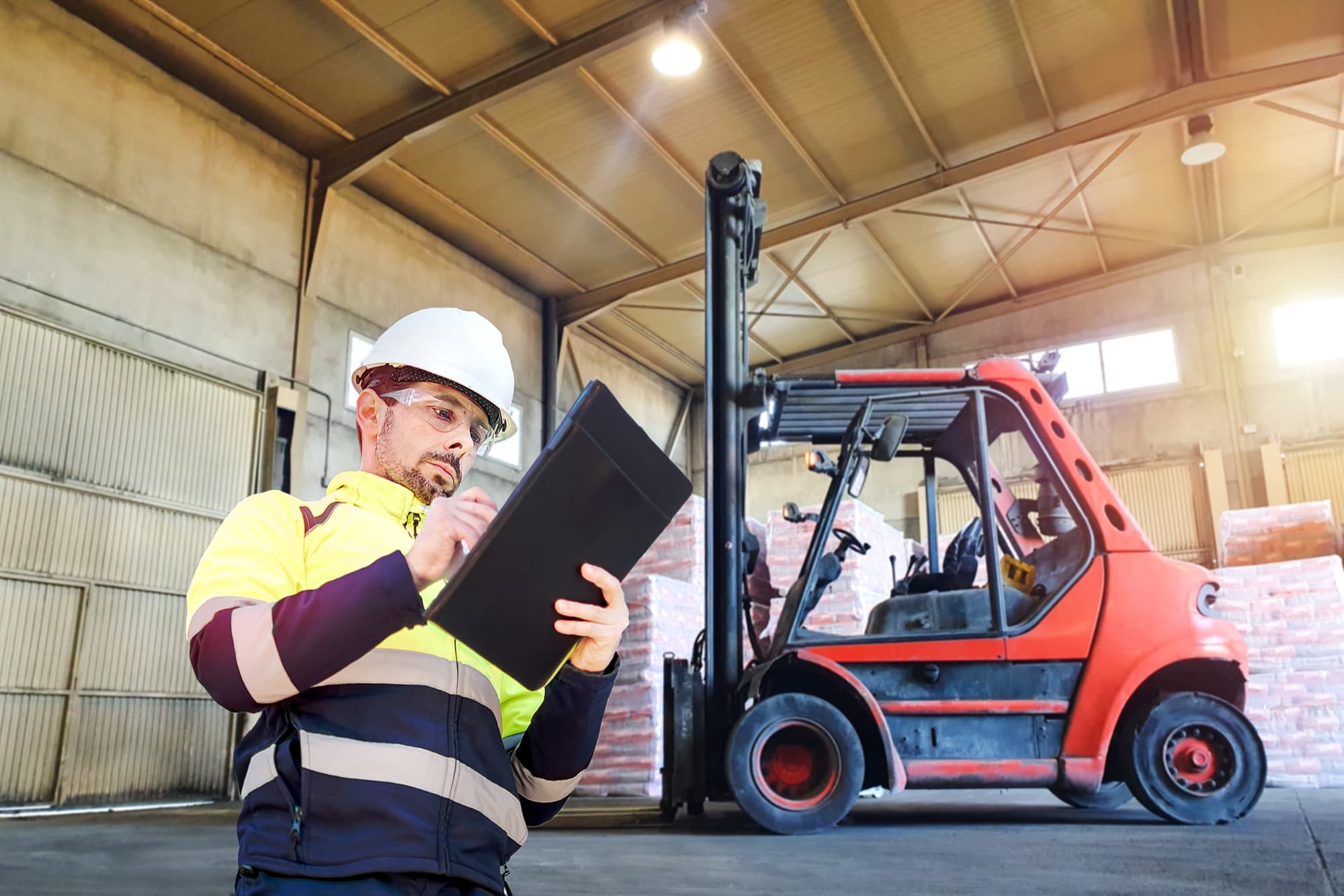 A worker in a hard hat, safety glasses, and a high-visibility jacket holds a clipboard in a warehouse, with a red forklift and stacked pallets in the background.