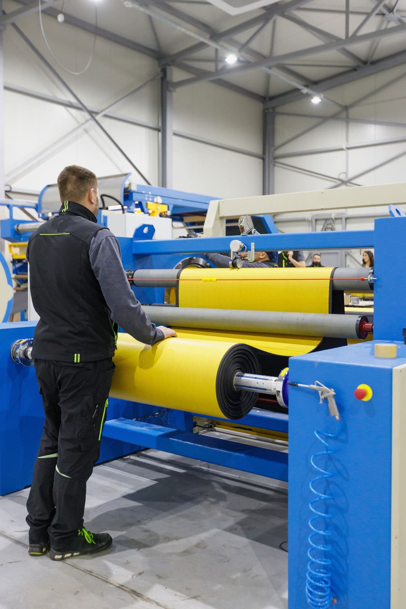 Operator with one hand on yellow roll material passing through rollers on industrial production equipment.