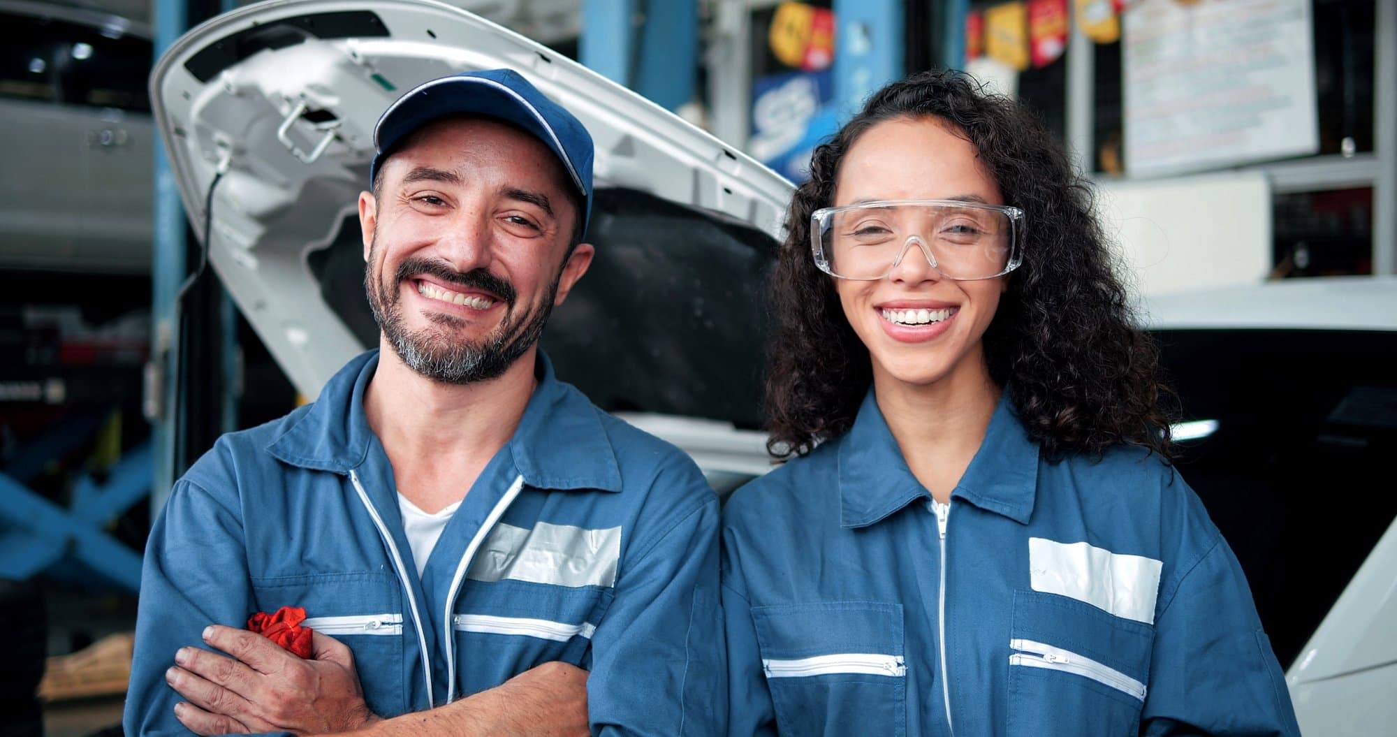 Two automotive service workers standing in front of a vehicle with its hood open.