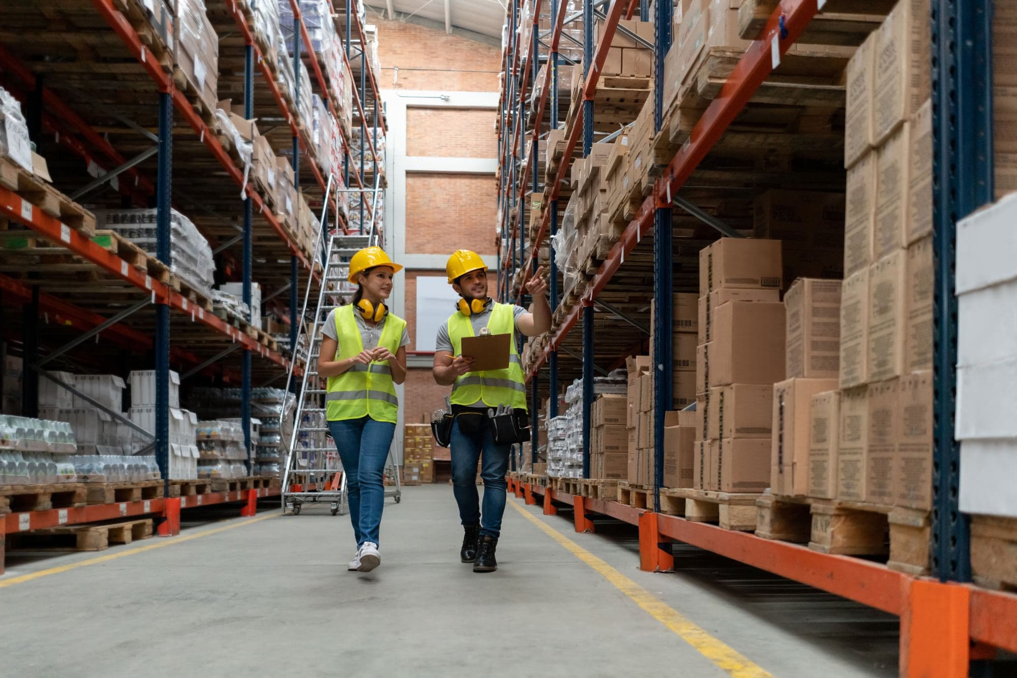 Two workers wearing hard hats and high-visibility vests walking through a warehouse aisle, one holding a clipboard and pointing toward stacked boxes on racking.