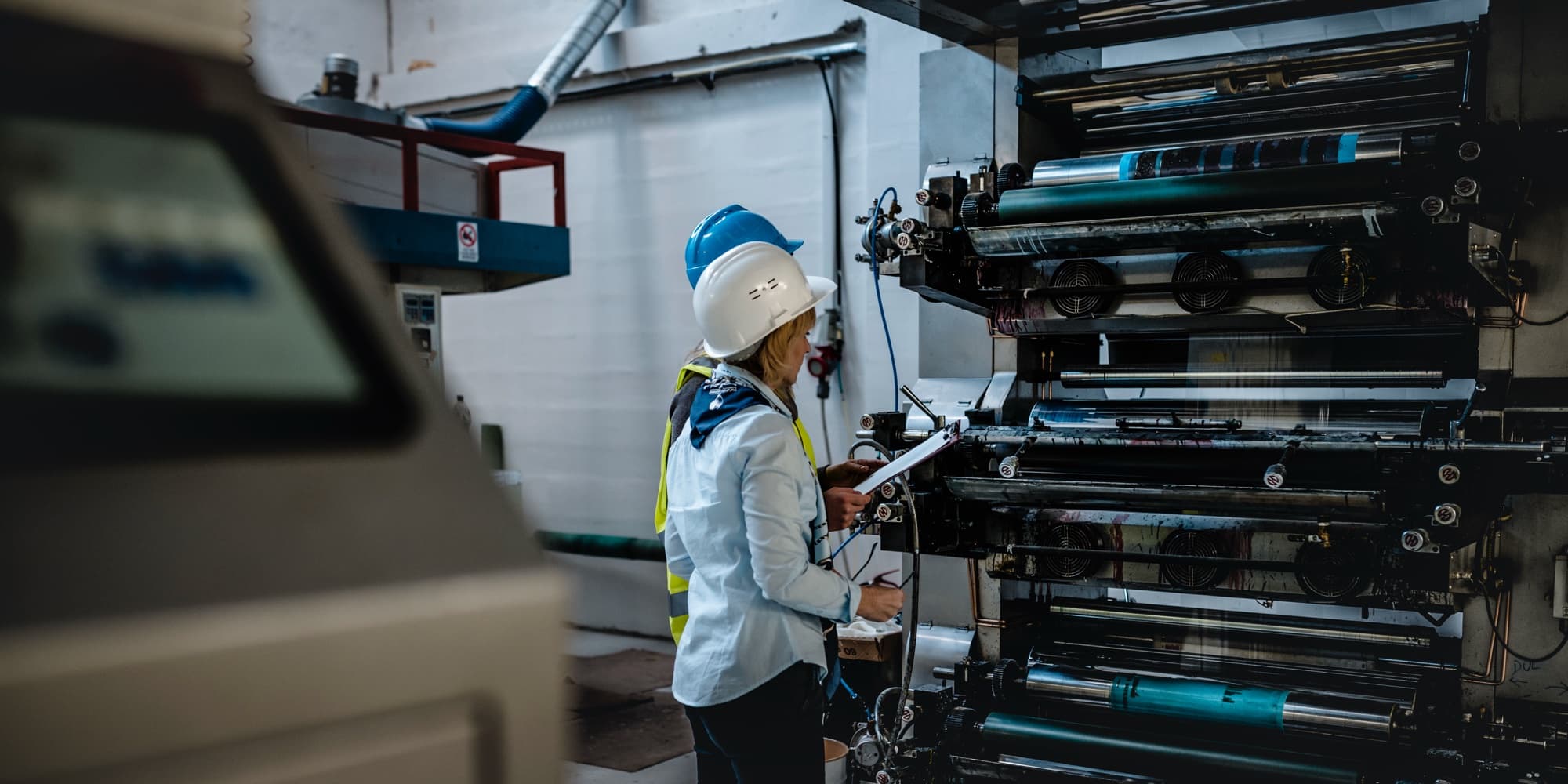 Worker in a white hard hat and high-visibility yellow vest standing beside a large multi-unit printing press with inked rollers, holding a clipboard