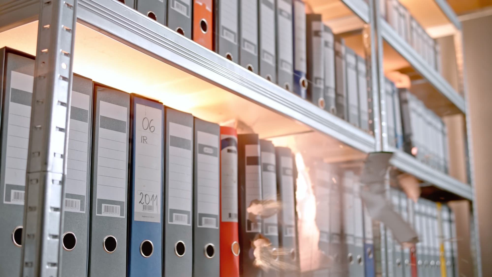 Rows of ring binders standing upright on metal shelving, most with light grey spines and a few in blue, red, and orange, some carrying handwritten labels