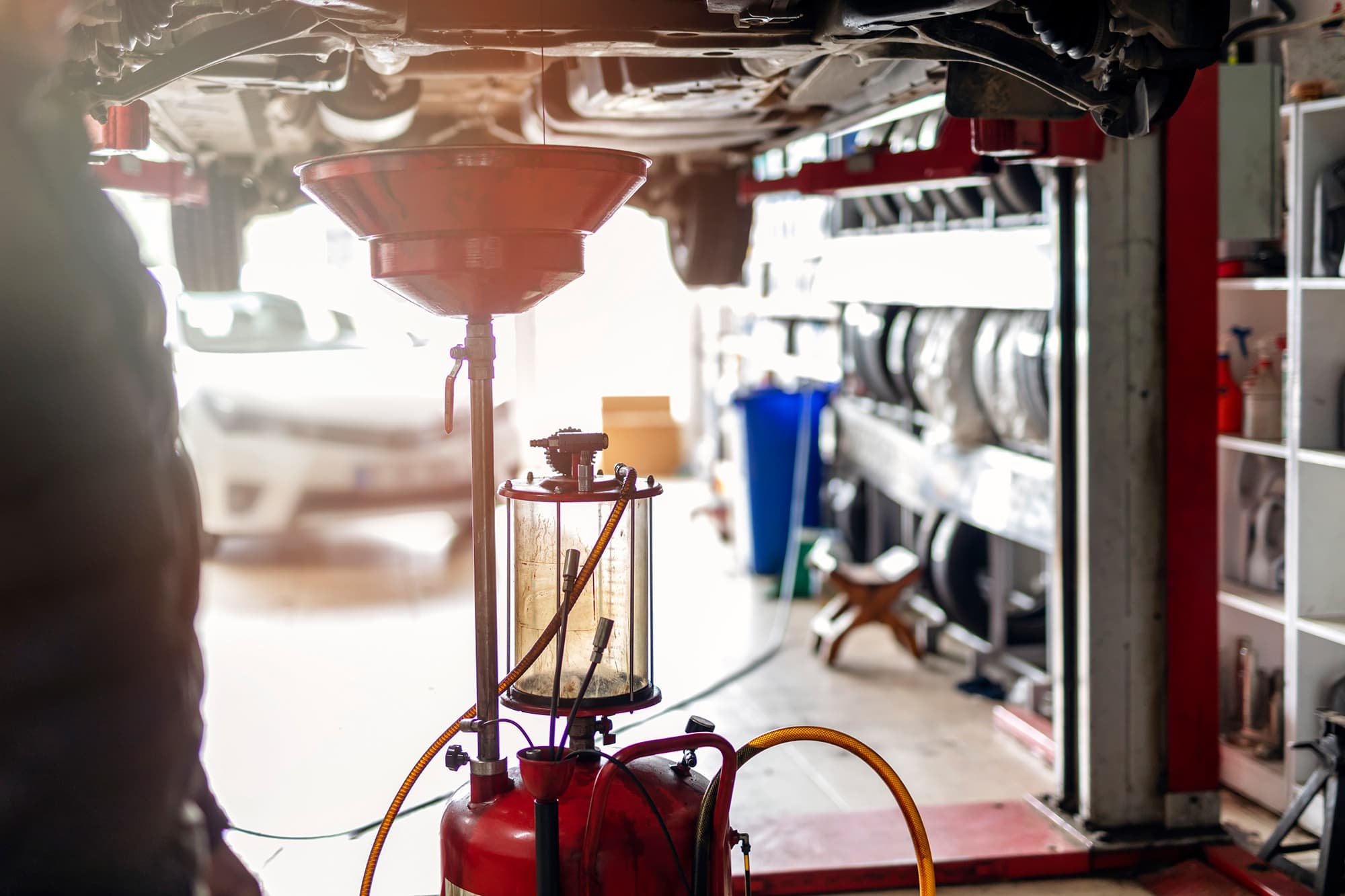 Red oil drain funnel on a stand catching fluid beneath a car raised on a lift, with tire racks and shelving in the background