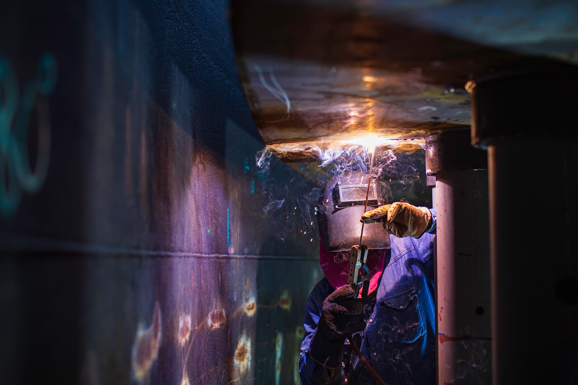 Welder in a protective hood working overhead inside a dark steel tank, with smoke and sparks lit by the welding arc