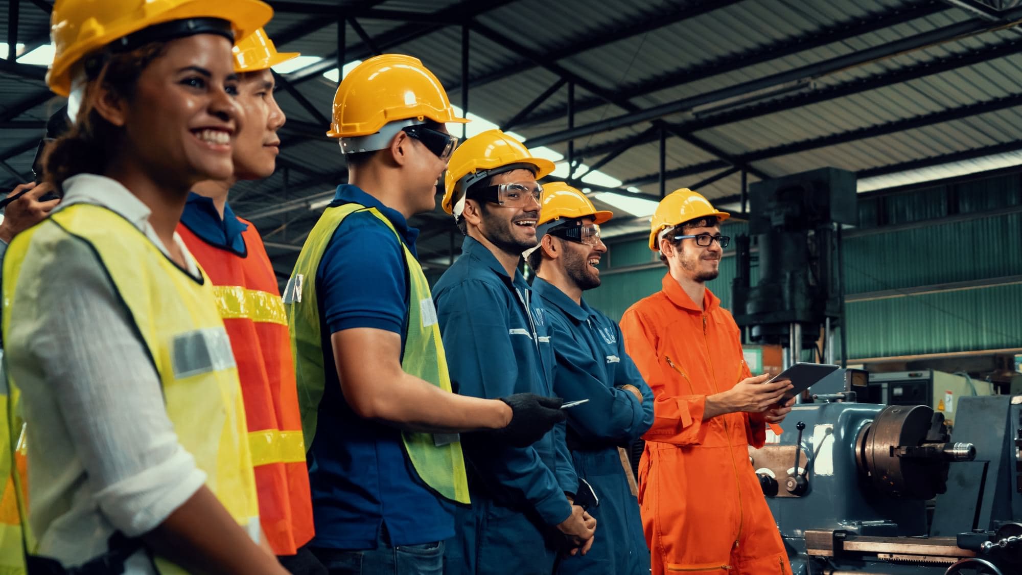 Group of workers wearing hard hats, high-visibility vests, and coveralls standing together in an industrial facility, with one holding a tablet.