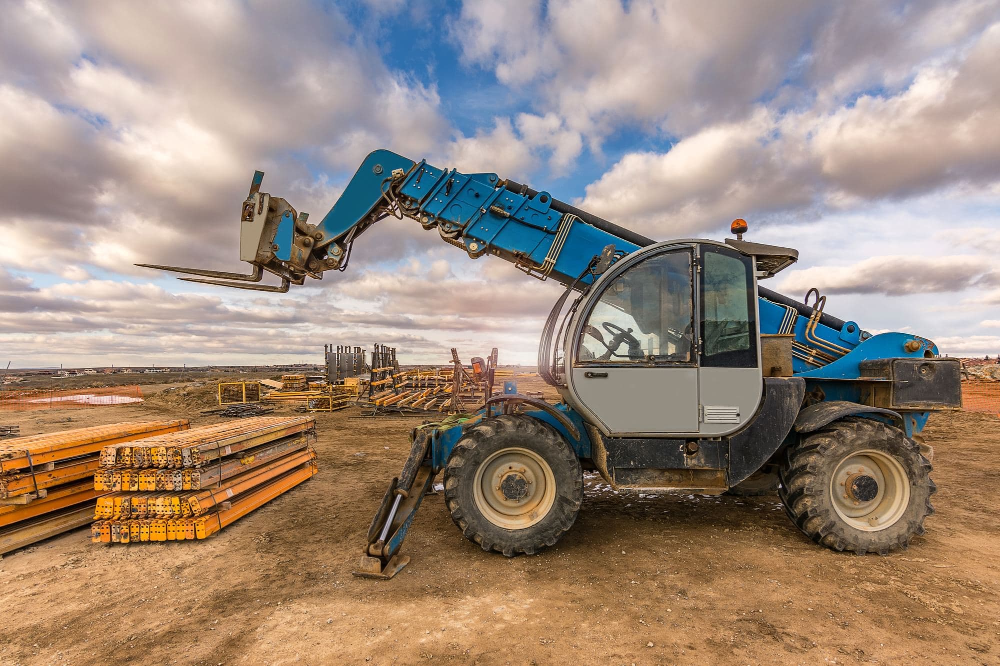 Blue telehandler with its boom raised and forks attached, parked on bare dirt at a construction site beside stacks of orange steel materials under a partly cloudy sky