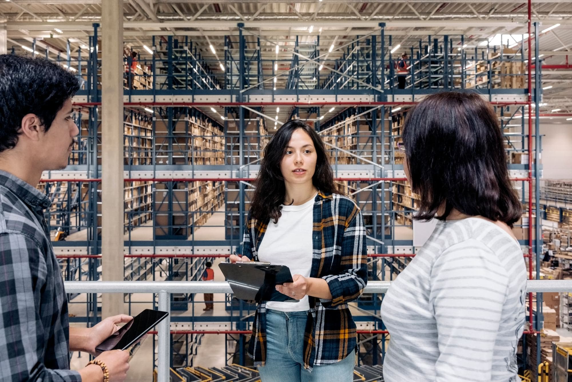 Three warehouse workers with a clipboard and a tablet talking on an elevated walkway in front of tall pallet racking.