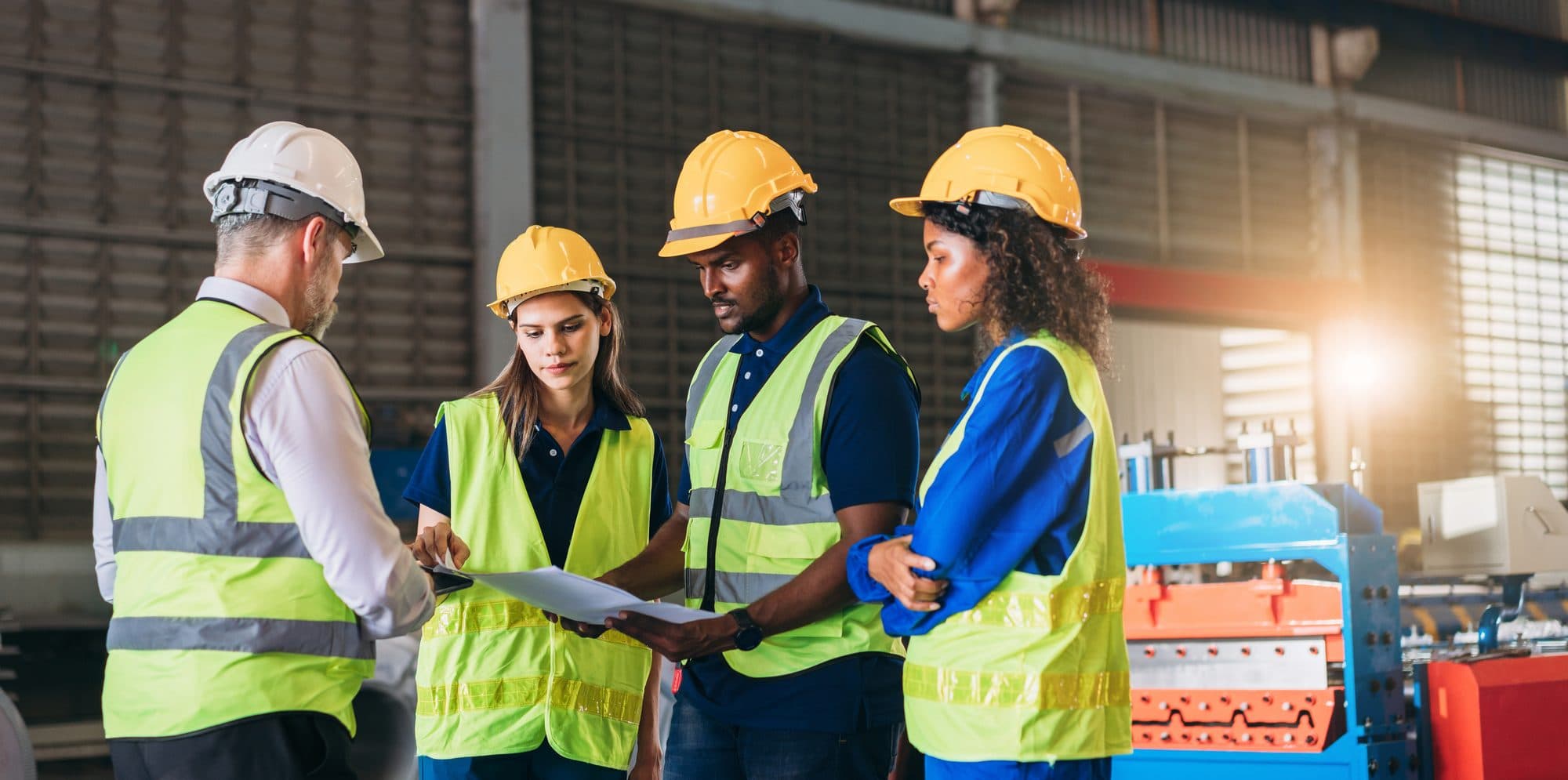 Four workers in high-visibility vests and hard hats reviewing a document together on an industrial plant floor, with rolling machinery behind them