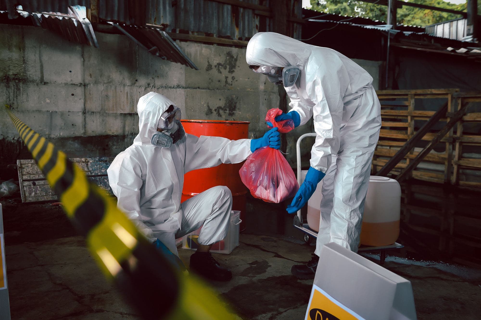 Two workers in hooded protective suits and respirators handling a red hazardous waste bag beside an orange drum