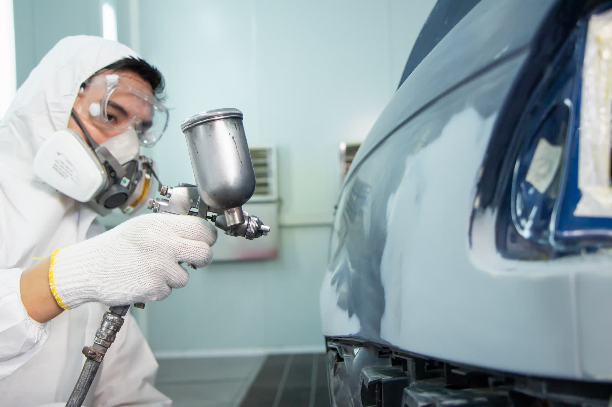 Trabajador con respirador y gafas de protección pintando con pistola el panel blanco de un vehículo en una cabina de pintura